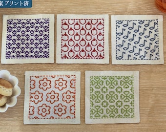 May include: Four square fabric coasters with different embroidered designs. The coasters are white with a light brown border. The designs are: purple geometric pattern, red geometric pattern, blue musical notes, orange floral pattern, and green geometric pattern.