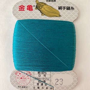 May include: A spool of teal-colored silk thread with a white label that reads "絹100% 9号 23dtex×9×2(s) 80m巻 普通染 金亀糸業(株) TEL 03-5687-8511" and "23". The label also features a turtle graphic and the text "金亀 登録商標 絹手縫糸".
