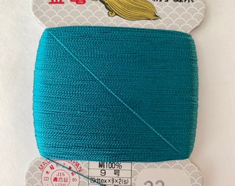 May include: A spool of teal silk thread with a white label that reads "絹100% 9号 23dtex×9×2(s) 80m巻 普通染 金亀糸業(株) TEL 03-5687-8511" and "23". The label also features a turtle graphic and the text "金亀 登録商標 絹手縫糸".