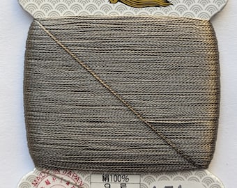 Puede incluir: Una bobina de hilo de seda 100% en color gris oscuro. La bobina está etiquetada con "金亀" (Kin-Kame) y "絹手縫糸" (Kinute Nuiito) en japonés. La etiqueta también incluye el texto "JIS 適合証 L2310 QTEC" y "再生紙使用" (Saisei Shi  Shiyou). La bobina está etiquetada con el número "151" y el texto "絹100% 9号 23utex×9×2(s) 80m巻 普通染 金亀糸業(株) TEL 03-5687-8511".