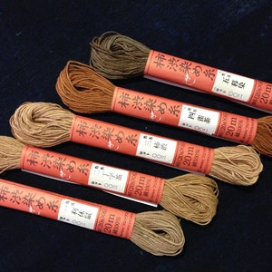 May include: Six spools of brown cotton thread, each wrapped in a red label with Japanese text and the words "柿渋染め糸" (Kakishibu Some Ito) which translates to "Persimmon Dye Thread". Each spool is labeled with the thread color, the length (20m), and the material (cotton).