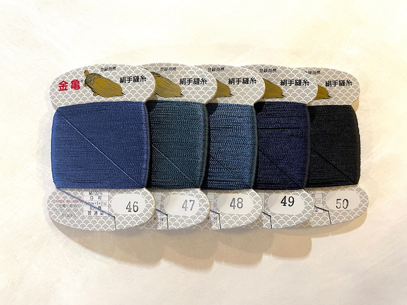 May include: Five spools of thread in shades of blue and black, labeled with numbers 46 through 50. Each spool is rectangular and wrapped in a paper label with Japanese text and a gold logo. The thread is likely for sewing or embroidery.