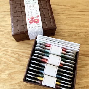 May include: A brown box with a woven pattern and a label that reads "さぬいと たまてばこ kimuito-tamatebako 冬色"  The box is open and contains a set of 10 spools of embroidery floss, each wrapped in a small, white paper packet. The spools are arranged in a row, and each one is a different color.