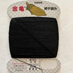 May include: A spool of black sewing thread with a paper label. The label features Japanese text, a gold turtle illustration, and the text "MADE IN JAPAN". The thread is 80 meters long.