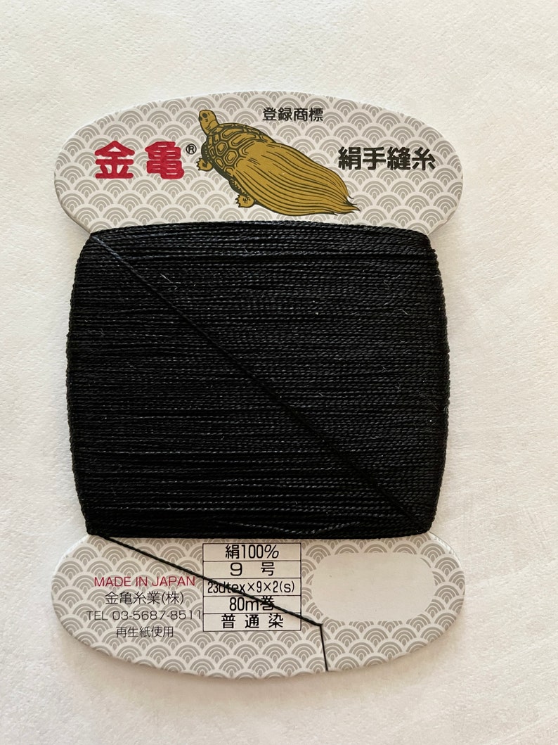 May include: A spool of black sewing thread with a paper label. The label features Japanese text, a gold turtle illustration, and the text "MADE IN JAPAN". The thread is 80 meters long.
