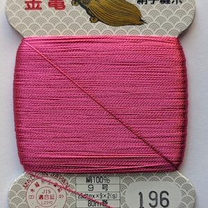 May include: A spool of pink thread with a label that reads "絹手縫糸" (silk hand sewing thread) and "金亀" (Kin-Kame) in gold lettering. The label also includes the text "JIS 適合証 L2310 OTEC" and "再生紙使用" (recycled paper used). The spool number is 196.