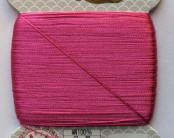 May include: A spool of pink thread with a label that reads "絹手縫糸" (silk hand sewing thread) and "金亀" (Kin-Kame) in gold lettering. The label also includes the text "JIS 適合証 L2310 OTEC" and "再生紙使用" (recycled paper used). The spool number is 196.