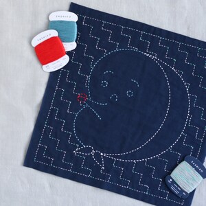 Puede incluir: Un cuadrado de tela azul con un dise&ntilde;o cosido blanco de una luna con cara y una flor. Cuatro carretes de hilo de bordar en rojo, blanco, azul y verde est&aacute;n sobre una superficie blanca.