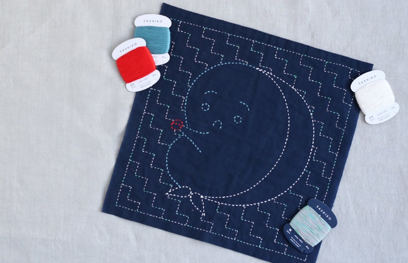 Puede incluir: Un cuadrado de tela azul con un dise&ntilde;o cosido blanco de una luna con cara y una flor. Cuatro carretes de hilo de bordar en rojo, blanco, azul y verde est&aacute;n sobre una superficie blanca.