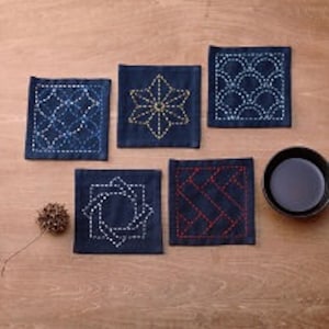 May include: Set of five blue fabric coasters with embroidered geometric designs in white and red thread. The coasters are square and have a subtle sheen.