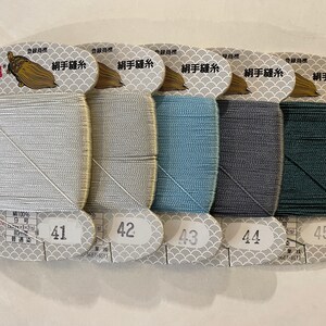 May include: Five spools of silk sewing thread in shades of silver, light blue, blue, gray, and dark green. Each spool is labeled with a number and has Japanese text. The thread is wound on a white card with a decorative pattern.