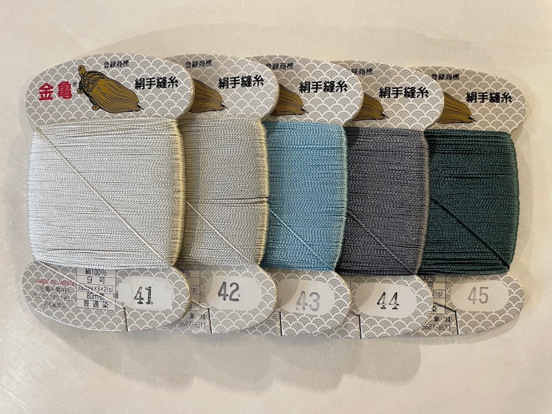 May include: Five spools of silk sewing thread in shades of silver, light blue, blue, gray, and dark green. Each spool is labeled with a number and has Japanese text. The thread is wound on a white card with a decorative pattern.