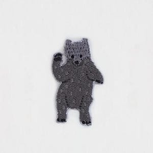May include: A grey embroidered iron-on patch of a bear waving. The bear is standing on its hind legs with its arms raised.