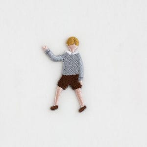 May include: Embroidered iron-on patch of a person with blonde hair wearing a gray sweater and brown shorts.
