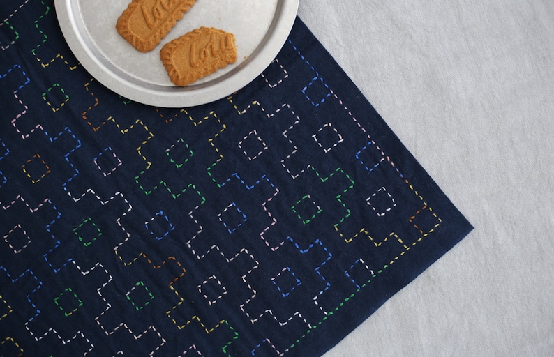 Puede incluir: Un mantel individual de tela azul oscuro con un patr&oacute;n geom&eacute;trico de costuras de colores. El mantel individual est&aacute; sobre una superficie blanca con dos galletas en un plato de plata.