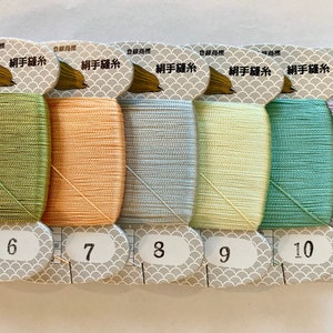 May include: A collection of six spools of colorful sewing thread. The threads are in shades of green, peach, light blue, yellow, teal, and lavender. Each spool is labeled with a number from 6 to 11. The packaging includes Japanese text and a gold acorn logo.