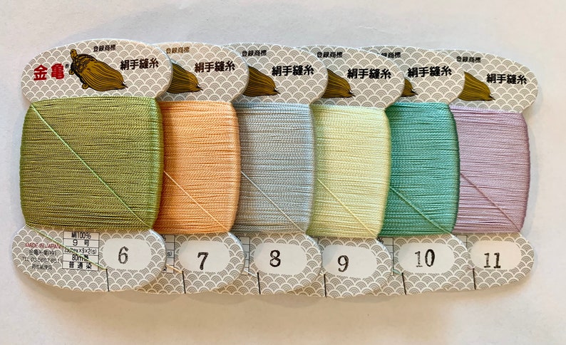 May include: A collection of six spools of colorful sewing thread. The threads are in shades of green, peach, light blue, yellow, teal, and lavender. Each spool is labeled with a number from 6 to 11. The packaging includes Japanese text and a gold acorn logo.