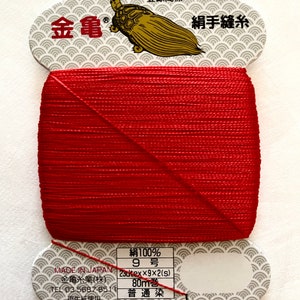 May include: A spool of red silk thread with a label that reads "絹100% 9号 230tex×9×2(s) 80m巻 普通染" and "金亀" in gold lettering. The label also features a turtle illustration.