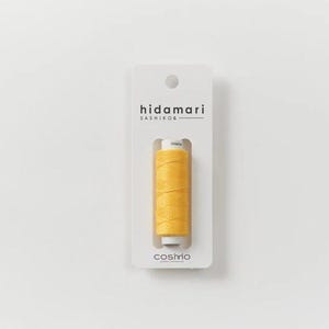 May include: A spool of bright yellow thread for sashiko embroidery, mounted on a white rectangular cardboard display card. The card features the text hidamari SASHIKO and cosmo at the bottom, with a circular hole at the top for hanging.