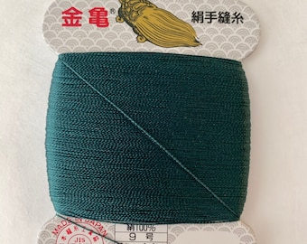 May include: A spool of dark green silk thread with a label that reads "金亀" (Kin-Kame) and "絹手縫糸" (Kinute Nuiito) in Japanese. The label also includes information about the thread, such as the material, size, and length. The label is decorated with a stylised turtle.