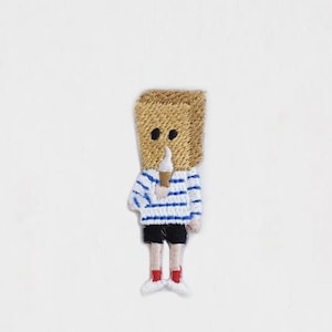 May include: Embroidered patch of a person with a brown paper bag over their head, wearing a blue and white striped shirt, black shorts, and red shoes. The person is holding a white ice cream cone.