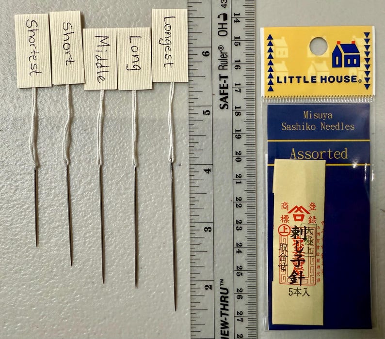 Pu&ograve; includere: Cinque aghi per sashiko di diverse lunghezze, etichettati "Shortest", "Short", "Middle", "Long" e "Longest". Gli aghi sono confezionati in un sacchetto di plastica trasparente con un'etichetta blu e bianca che recita "Misuya Sashiko Needles Assorted" e presenta una piccola icona di casa. L'etichetta include anche testo giapponese.