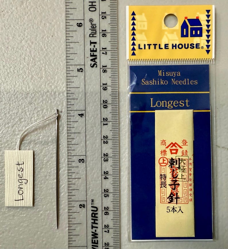 Pu&ograve; includere: Un pacchetto di aghi Sashiko Misuya etichettato "Longest". La confezione &egrave; gialla con testo nero e un sigillo rosso. Gli aghi sono esposti su una carta bianca con il testo "Longest" scritto su di essa.