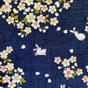 May include: Dark blue fabric with a pattern of white rabbits, pink and white cherry blossoms, and falling petals. The blossoms have yellow centers and green leaves. The fabric has a textured appearance.