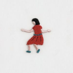 May include: Embroidered patch featuring a figure in a red dress with a blue belt and shoes, arms outstretched. The figure has black hair and is in a dancing pose. Ideal for clothing or accessories.