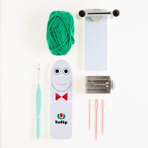 May include: A white plastic loom with a smiling face, red bow tie, and the word "Tulip" printed on it. The loom is accompanied by a ball of green yarn, a crochet hook, a metal yarn winder, and three pink knitting needles.