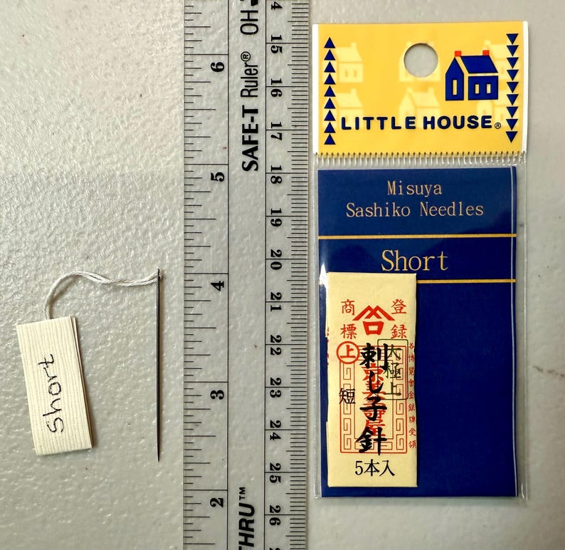 Pu&ograve; includere: Un pacchetto di aghi sashiko corti di Misuya, etichettato "Little House" e "Short". La confezione &egrave; blu con lettere bianche e una piccola illustrazione di una casa. Gli aghi sono confezionati in una piccola busta bianca con testo giapponese.