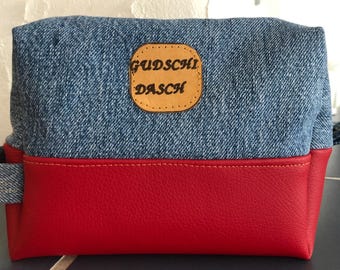 May include: A rectangular denim and red pouch with a leather patch that reads "Gudschi Dasch". The top half is blue denim, and the bottom half is red. The pouch has a small handle on the side.