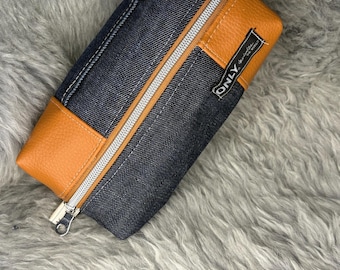May include: A rectangular denim pencil case with a brown leather accent and a silver zipper. The case has a tag that says "ONLY" and another tag that says "love". The case is on a gray, furry surface.