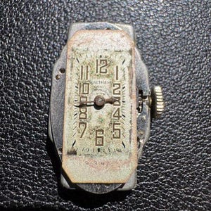 Op de afbeelding: Een zilverkleurig Waltham-horloge met zwarte Romeinse cijfers voor de uren en zwarte Arabische cijfers voor de minuten. De wijzerplaat is ovaalvormig en heeft een witte achtergrond. De wijzerplaat is gemarkeerd met "Waltham" en "913 RF".