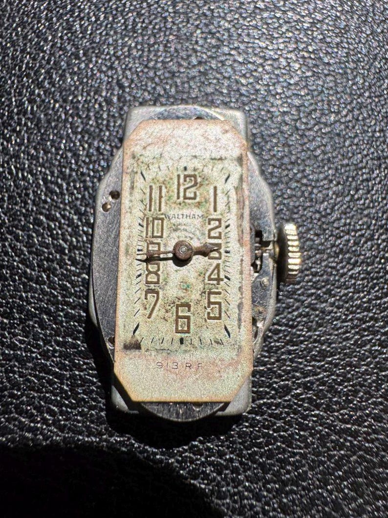 Op de afbeelding: Een zilverkleurig Waltham-horloge met zwarte Romeinse cijfers voor de uren en zwarte Arabische cijfers voor de minuten. De wijzerplaat is ovaalvormig en heeft een witte achtergrond. De wijzerplaat is gemarkeerd met "Waltham" en "913 RF".