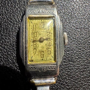 Op de afbeelding: Een zilveren polshorloge met een rechthoekige wijzerplaat en een gouden wijzerplaat. Het horloge heeft een zwarte leren band en een witte wijzerplaat met zwarte Romeinse cijfers als uurmarkeringen. De wijzerplaat van het horloge heeft ook een kleine secondewijzer.