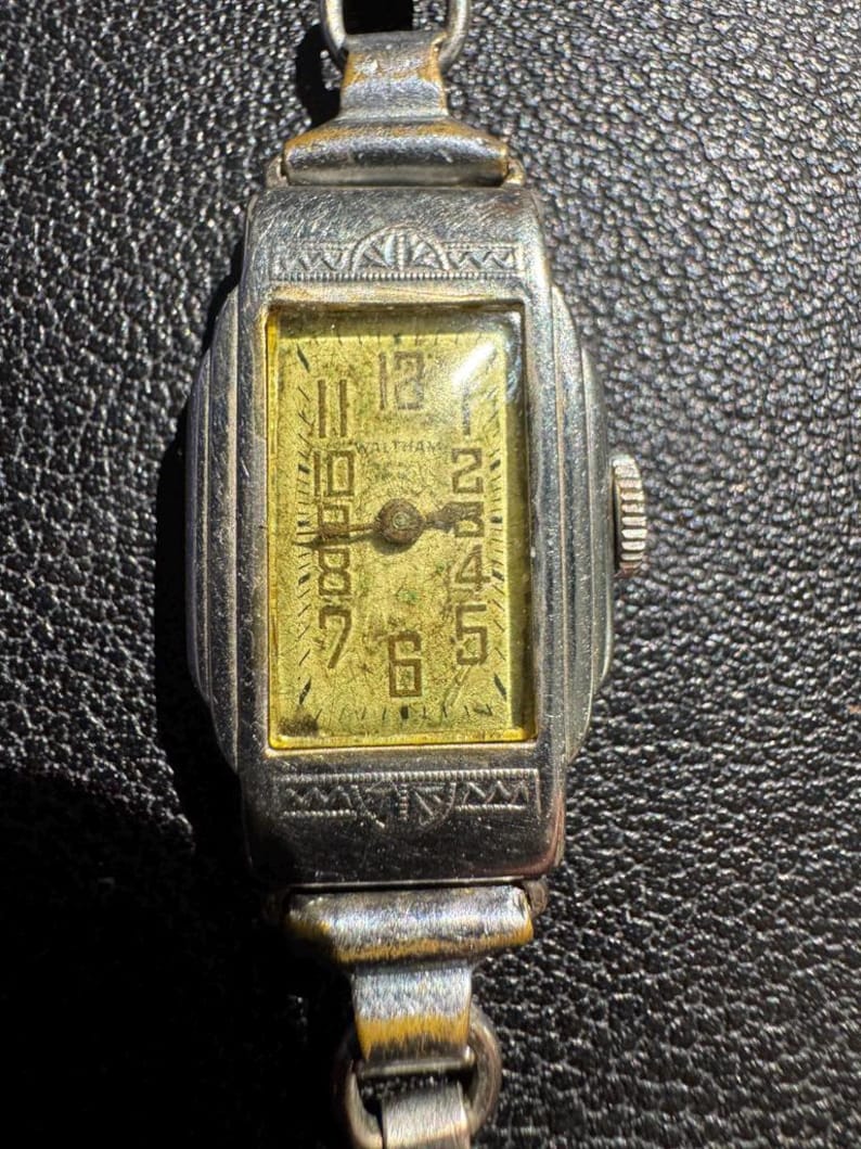Op de afbeelding: Een zilveren polshorloge met een rechthoekige wijzerplaat en een gouden wijzerplaat. Het horloge heeft een zwarte leren band en een witte wijzerplaat met zwarte Romeinse cijfers als uurmarkeringen. De wijzerplaat van het horloge heeft ook een kleine secondewijzer.