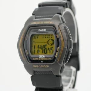 May include: A black Casio digital watch with a yellow-tinted display screen. The watch face features the text Illuminator, Casio, 10 Year Battery, HD, Alarm Chronograph, and WR 100M. The digital display shows the time 11:46:45 and a sun icon.