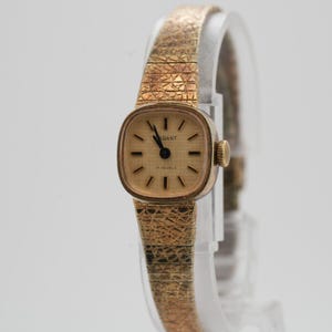 May include: A gold-coloured vintage wristwatch with a textured strap and a square face. The watch face displays the word "ELEGANT" and the words "17 JEWELS". The watch has a small, round crown. The watch face is light-coloured with black hands and markers.
