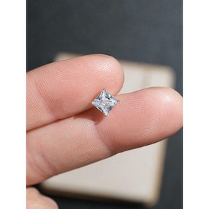 Peut inclure: Un petit bijou carré et transparent, peut-être un diamant ou une zircone cubique, serti dans un métal argenté ou blanc. La pierre précieuse est tenue entre deux doigts, mettant en valeur ses facettes et son éclat. L'arrière-plan est flou et neutre.