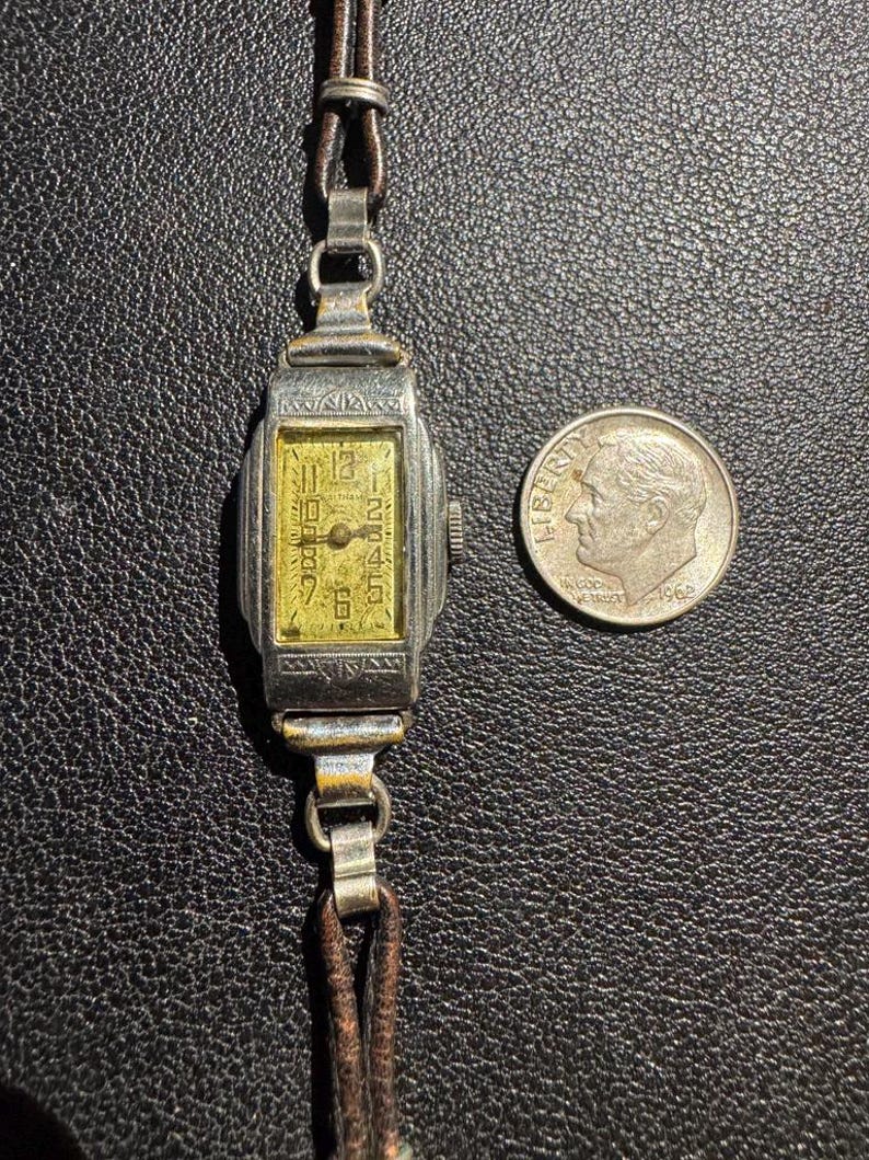 Op de afbeelding: Een zilveren polshorloge met een rechthoekige wijzerplaat en een bruin leren bandje. De wijzerplaat van het horloge heeft een gele achtergrond met zwarte Romeinse cijfers als uurmarkeringen en zwarte wijzers. Het horloge is ongeveer zo groot als een Nederlandse tien-cent munt.