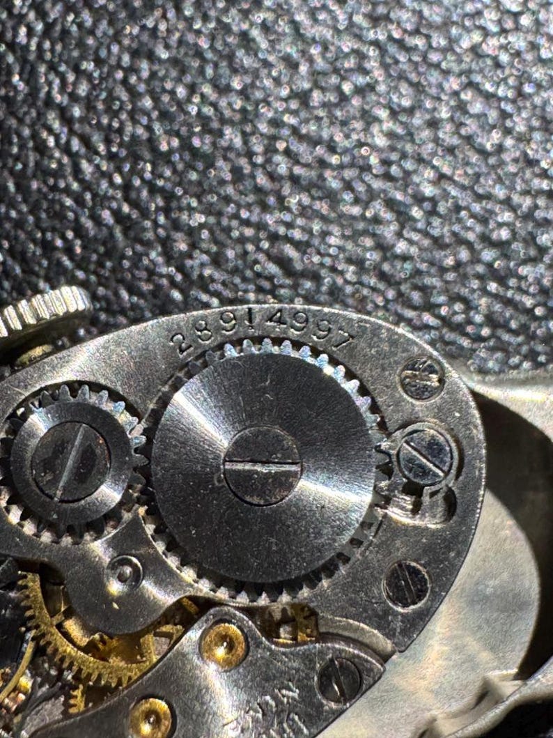 Op de afbeelding: Close-up van een zilveren uurwerkmechanisme met zichtbare tandwielen en de gegraveerde nummers 2891 4997 op de buitenring.