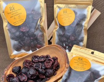 Op de afbeelding: Drie zakken met gevriesdroogde biologische rode kersen. De zakken zijn goudkleurig met doorzichtige plastic ramen die de kersen laten zien. De zakken hebben een etiket met de tekst "Rk Freeze dried goodies ORGANIC RED CHERRIES KEEP IT ZIPPED KEEP IT CRISP".