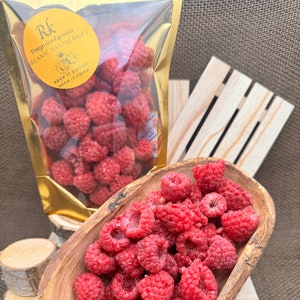 May include: A bowl of freeze-dried organic raspberries. The raspberries are a vibrant red color and are piled high in the bowl. The bowl is made of wood and has a natural finish. The raspberries are packaged in a gold bag with the text "Freeze dried goodies Organic Raspberries Keep it zipped Keep it crisp".