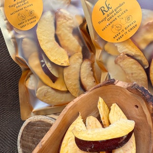 May include: Two clear plastic bags with gold accents, each containing dried nectarine slices. The bags have a yellow circle label with the text "Rk Freeze dried goodies ORGANIC NECTARINE SLICES KEEP IT ZIPPED KEEP IT CRISP". A wooden bowl filled with dried nectarine slices.