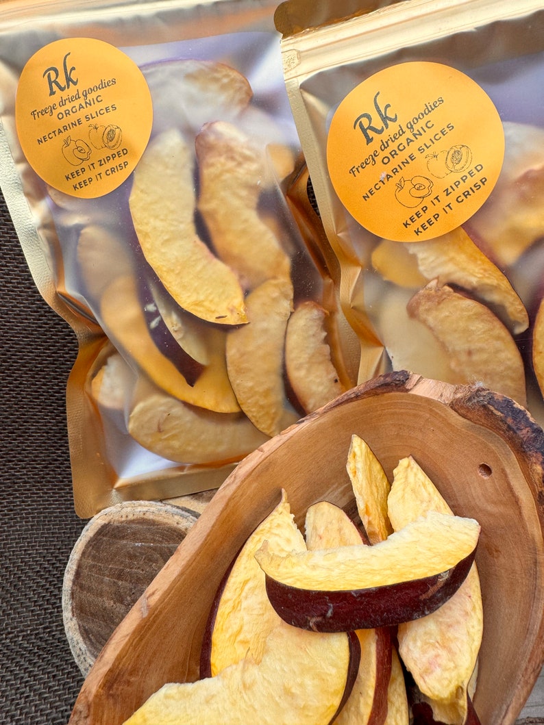May include: Two clear plastic bags with gold accents, each containing dried nectarine slices. The bags have a yellow circle label with the text "Rk Freeze dried goodies ORGANIC NECTARINE SLICES KEEP IT ZIPPED KEEP IT CRISP". A wooden bowl filled with dried nectarine slices.