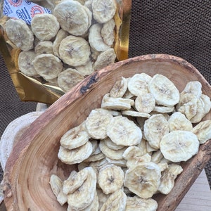 May include: A bowl of dried banana chips. The chips are light brown and have a slightly translucent appearance. The bowl is made of wood and has a natural finish. The chips are in a bag with the text "Keep it zipped, keep it crisp".