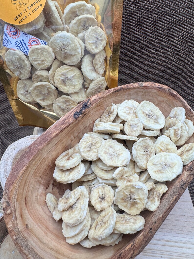 May include: A bowl of dried banana chips. The chips are light brown and have a slightly translucent appearance. The bowl is made of wood and has a natural finish. The chips are in a bag with the text "Keep it zipped, keep it crisp".