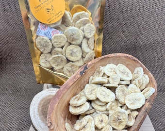 May include: A gold bag with a zipper closure contains freeze-dried organic banana slices. The bag has a label that reads "Freeze dried goodies, Organic Bananas, Keep it zipped, Keep it crisp". A wooden bowl filled with banana slices sits on a wooden surface.