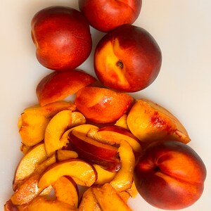 May include: A white cutting board with four whole nectarines and several slices of nectarine. The nectarines are red and orange with a yellow center. The slices are yellow and orange.
